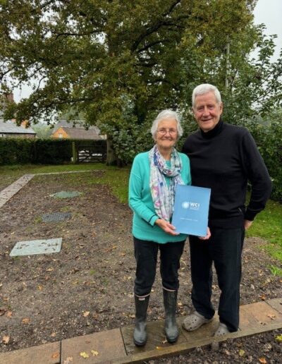 A couple posing in front of a newly installed sewage treatment plant with a presentation folder containing a cheque